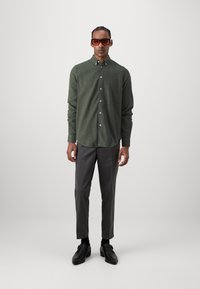Dark green button-up shirt with long sleeves, paired with gray trousers and black shoes. The shirt has a slight texture and rounded hem.
