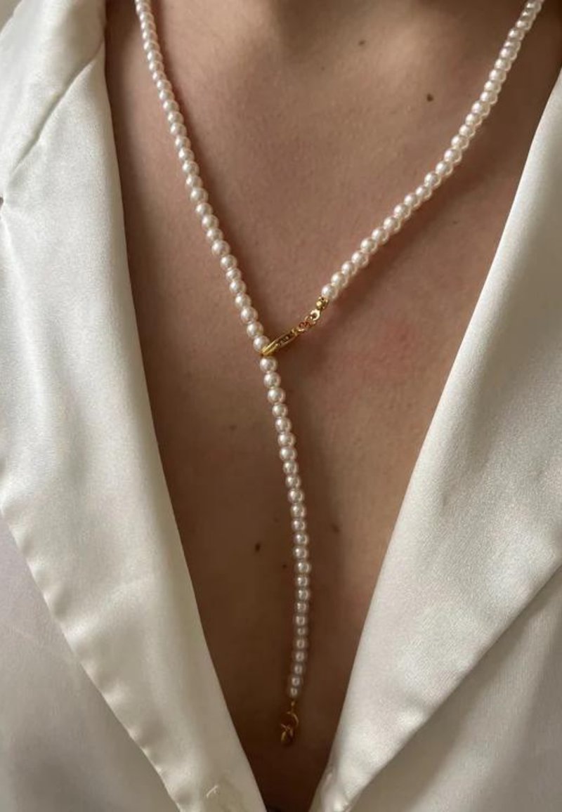Gros plan d’un collier de perles avec fermoir en or porté sur une peau nue sous un chemisier blanc en satin avec un décolleté profond.
