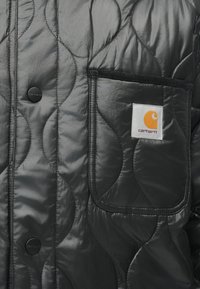 Czarna pikowana kurtka z kieszenią naszywaną, gładką teksturą i metką Carhartt. Zapięcie na napy oraz wygięty wzór pikowania.