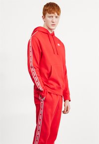 Rdeča kapuca in trenirka iz mehkega blaga. Kapuca ima sprednji žep in logotip; hlače imajo bele stranske črte z blagovno znamko.