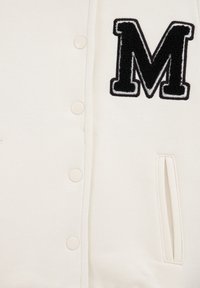 Veste universitaire off-white avec un patch "M" texturé noir, boutons-pression et poches latérales. Tissu doux avec une texture lisse.