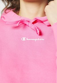 Felpa rosa con cappuccio e coulisse. Presenta un logo "Champion" bianco. Il tessuto sembra liscio e leggermente texture.