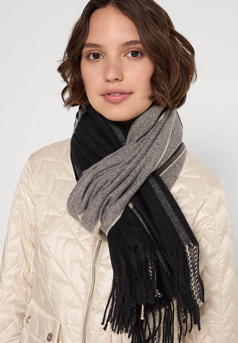 Czarny i ciemnoszary szalik w paski wykonany z miękkiego materiału, ozdobiony frędzlami. Swobodnie zawiązany wokół szyi, na jasnej, pikowanej kurtce.
