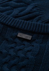 Merinvärd kabelstickad tröja med en texturerad design och en svart rektangulär etikett med "QUINTESSENCE" präglad på den.