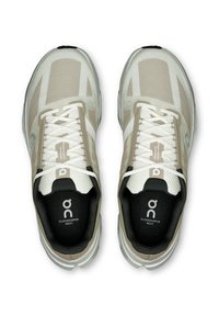 Baskets de course beige clair avec une tige en mesh, accents blancs et semelle texturée. Présente un logo prononcé et un système de laçage détaillé.