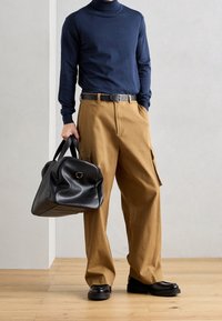 Marinefarbener Rollkragenpullover, beigefarbene Cargo-Hosen mit Taschendetails, schwarze Leder-Duffel-Tasche und schwarze Schuhe mit dicker Sohle.