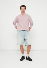 Mladý muž stojí opretý o bielu stenu, oblečený v ružovej mikine s dlhým rukávom, svetlých rifľových šortkách, bielych ponožkách a čiernych mokasínach.