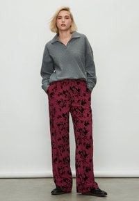 Bluză polo gri cu mâneci lungi și guler; pantaloni largi roz cu model și design floral negru; modelul poartă pantofi negri.
