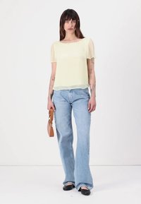Blouse légère jaune clair et transparente à manches courtes, design superposé, portée avec un jean en denim bleu taille haute, tenant un petit sac à main marron.