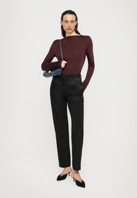 Woman stands wearing a maroon long-sleeve top, black pants, black heels, and holds a navy blue handbag with a silver chain strap.