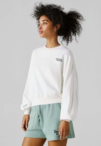 Ženska s kodrastimi lasmi, oblečena v belo majico s kapuco "Red Bull Racing" in svetlo zelene kratke hlače, stoji bočno ob sivem ozadju.