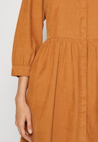 Robe en velours côtelé marron avec taille froncée et manches trois-quarts, montrant un gros plan du bas du torse et du bras gauche.