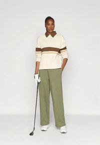 Roomkleurig en bruin gestreept poloshirt met een kraag, gecombineerd met olijfgroene broek met wijd uitlopende pijpen. Het model houdt een golfclub vast en draagt een handschoen.