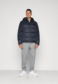 Marineblå pufferjakke med hætte, der har lynlås og quiltet design. Sammen med grå joggingbukser og hvide sneakers.