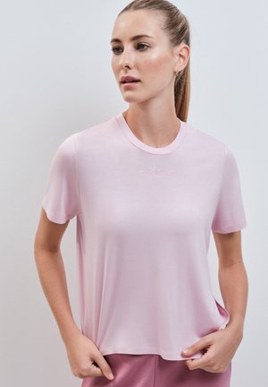 Femme aux longs cheveux portant un T-shirt rose clair à manches courtes et un pantalon rose foncé, debout, tenant l'ourlet du T-shirt, regardant sur le côté.