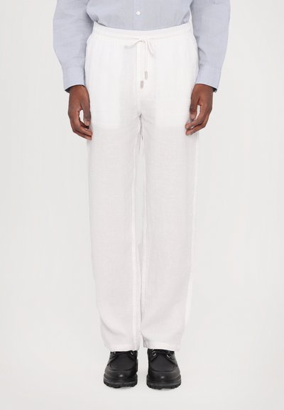 Pantalons en lin blanc avec une coupe décontractée, taille élastique à cordon de serrage et poches latérales, associés à des chaussures noires. Tissu texturé pour le confort.