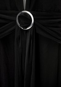 Vêtement en tissu noir avec un anneau central en métal argenté et des liens en tissu fluides. Matière texturée avec des détails de drapé doux.