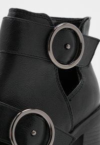 Musta nilkkuri, joka on valmistettu tekstuuripinnoitteisesta synteettisestä nahasta, jossa on kaksi hopeista pyöreää yksityiskohtaa ja avoin sivusuunnittelu. Näkyy pinoitu kantapää.