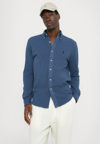 Chemise bleue à manches longues avec col, arborant un petit logo sur la poitrine et des boutons blancs, associée à un pantalon beige.