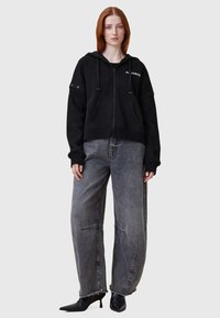 Sudadera negra con cremallera y cordones, grandes bolsillos frontales y acentos plateados; combinada con pantalones de mezclilla gris holgados y zapatos negros de punta.