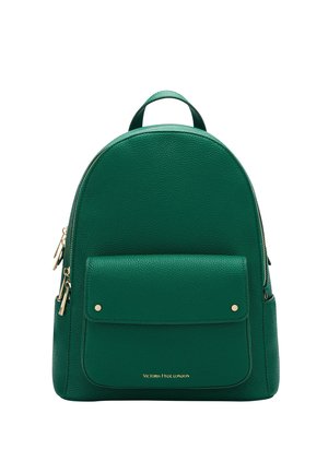 Osoba w beżowym płaszczu niesie zielony skórzany plecak z wieloma zamykanymi na zamek kieszeniami na zewnątrz w miejskim otoczeniu.