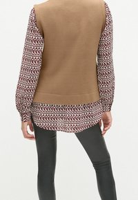 Brauner ärmelloser Weste über einer gemusterten burgunderfarbenen und weißen Bluse, kombiniert mit schwarzen Leggings, die strukturierte Stoffe und ein geschichtetes Design hervorhebt.