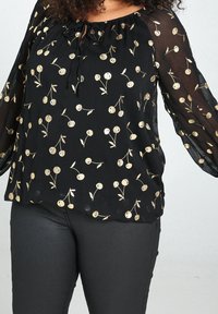 Blouse noire avec manches longues en voile transparent et col à cordon, ornée de motifs circulaires dorés et de petits dessins floraux.
