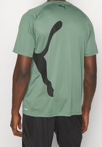 Zielony sportowy t-shirt z dużym czarnym logo na plecach, krótkimi rękawami, gładką fakturą i materiałem odprowadzającym wilgoć.