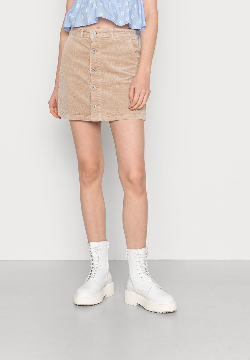 Saias mini de corduroy bege com fecho frontal em botão, apresentando bolsos e uma superfície texturizada, combinada com botas de combate brancas com atacadores.
