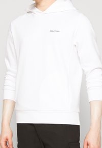 Witte katoenen hoodie met lange mouwen, voorzien van een minimalistisch ontwerp en het "Calvin Klein" logo in het zwart geborduurd op de borst.