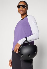 Lilla langermet skjorte med hvite ermer, kombinert med en svart hjelm holdt i hånden. Solbriller fullfører antrekket og fremhever den sporty stilen.