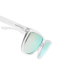 Prozorna, večja sončna očala z modro-zelenimi lečami z gradientom. Imajo prozoren okvir in kraka. Na strani je napisano blagovno ime "HAWKERS".