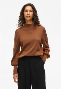 Pull en maille marron avec un col montant et des manches à motif texturé. Le corps est lisse, tandis que les manches présentent un motif en losange.