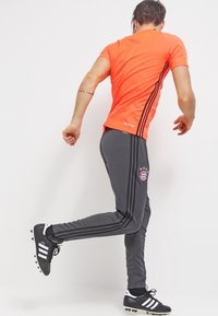 Oranje sportshirt met korte mouwen, gecombineerd met grijze trainingsbroek met zwarte strepen en een logo op de linkerdij, gedragen met zwarte schoenen.