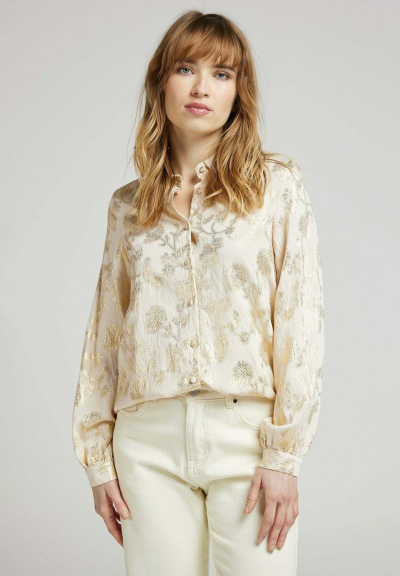 Chemise beige clair à boutons avec des motifs floraux dorés, manches longues avec des poignets à boutons, et coupe décontractée. Associée à un jean blanc.