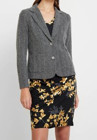 Šedý blazer ve vzoru herringbone s přiléhavým střihem, zapínáním na jeden knoflík a klopovými límci, v kombinaci s černými květinovými šaty s žlutými akcenty.