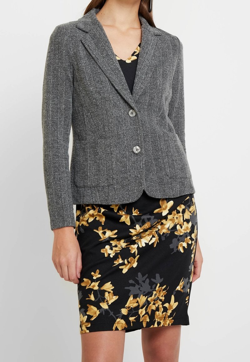 Šedý blazer ve vzoru herringbone s přiléhavým střihem, zapínáním na jeden knoflík a klopovými límci, v kombinaci s černými květinovými šaty s žlutými akcenty.