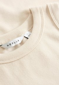 Gros plan sur un tissu en tricot côtelé de couleur crème, montrant le détail du col ras du cou avec une étiquette de marque NOELLA et une étiquette de taille petite.