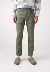 Mies, joka käyttää oliivinvihreitä slim-fit-housuja, valkoisia sukkia, beigen väreisiä liukuhihna-kenkiä ja valkoista collegepaitaa, jossa on sininen maisemakuva.