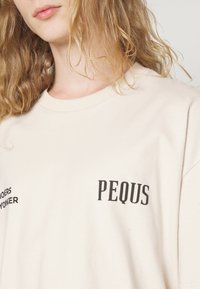 Beżowa bawełniana koszulka z krótkim rękawem, okrągłym dekoltem i czarnym napisem "PEQUS" wyraźnie umieszczonym na przodzie.
