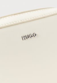 Kreemivärvi rahakott, sileda tekstuuriga, õmmeldud äärtega ja hõbedase "HUGO" logoga, mis on silmatorkavalt nähtav.