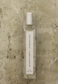 Bottiglia di profumo in vetro trasparente con un tappo cilindrico bianco. Presenta un'etichetta verticale bianca con testo nero, design minimalista e forma rettangolare.