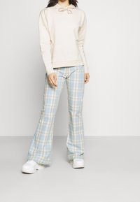 Sweat à capuche beige avec cordon de serrage, associé à un pantalon évasé à carreaux bleu clair. Le modèle porte des baskets blanches. Tissu doux, coupe décontractée.