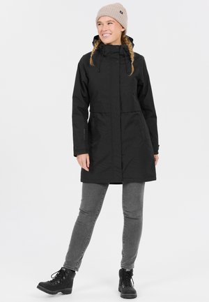 Svart vattentät parka med dragkedja och tryckknappslås, justerbar huva och framfickor. Figurvänlig design tillsammans med grå jeans och stövlar.