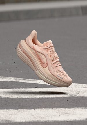 Chaussure de running Nike rose clair en suspension au-dessus d'une route asphaltée avec des lignes blanches peintes et de petits débris autour.