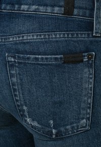 Jeans de mezclilla azul oscuro con una textura desgastada, que cuentan con un bolsillo trasero y una etiqueta negra encima. Diseño clásico de cinco bolsillos.