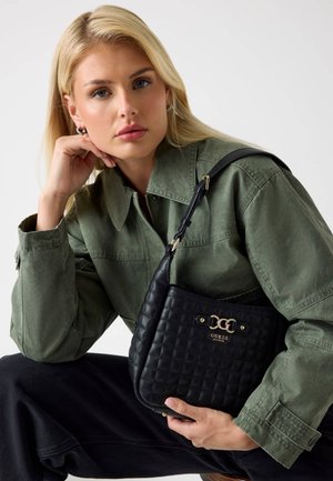 Sac à main matelassé noir avec quincaillerie dorée et logo. Le modèle porte une veste vert olive, mettant en valeur la bandoulière du sac.