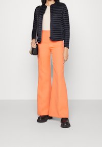 Navy puffer jacket with ribbed texture, paired with bright orange flared pants and chunky black footwear. Black crossbody bag accent.