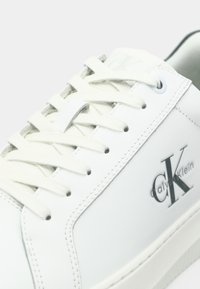 Baskets blanches Calvin Klein avec des lacets blancs, logo CK sur le côté et des coutures détaillées sur un fond clair.
