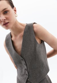 Grå herringbone-väst med breda armhål, djup V-hals och fem knappar. Har en liten ficka på vänster sida och textil med struktur.
